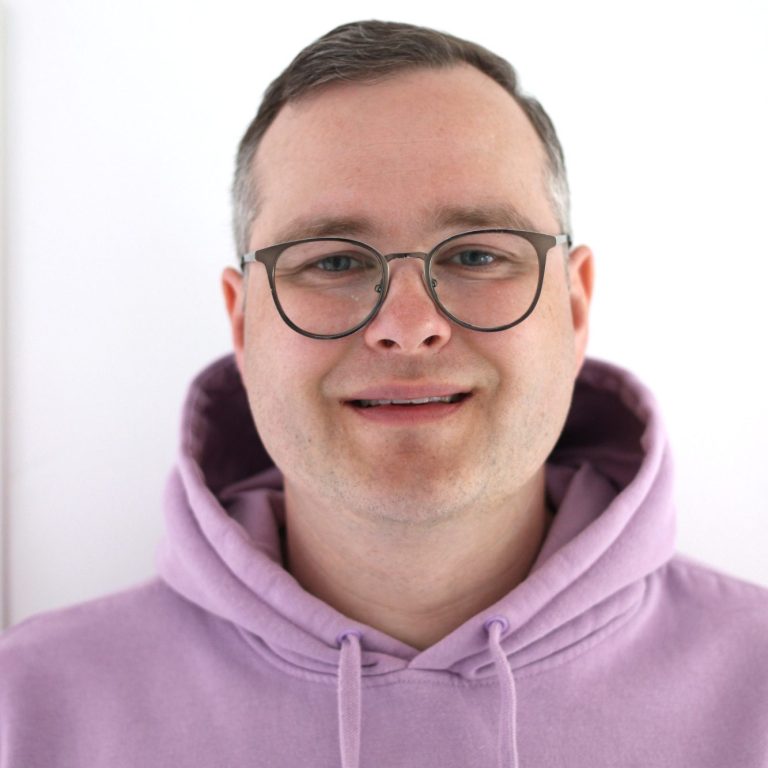 Lächelnder Mann mit Brille in lila Kapuzenpullover vor neutralem Hintergrund.