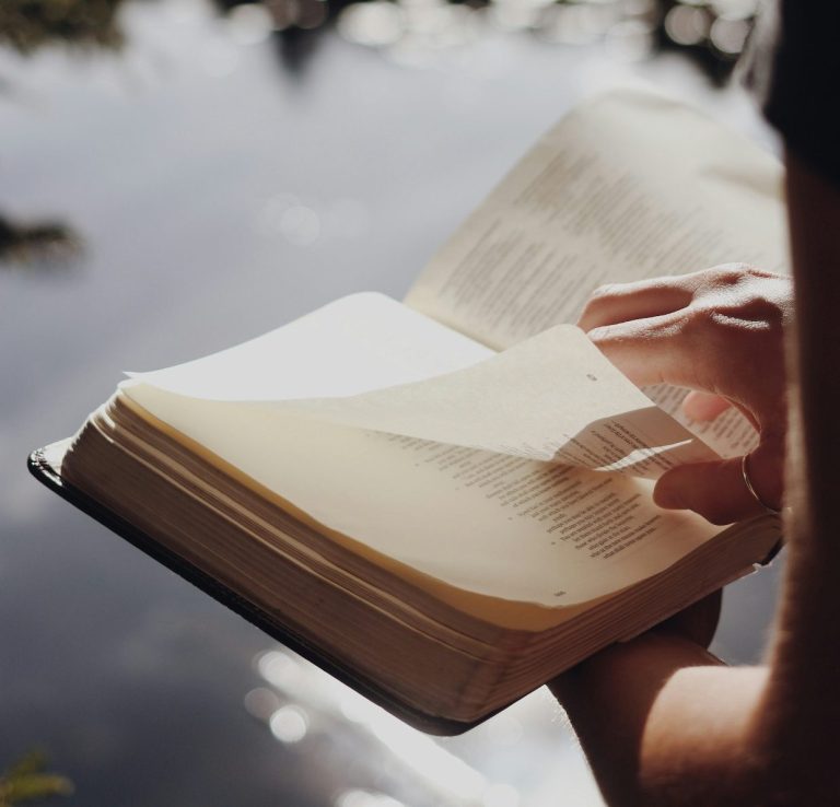 Eine Hand blättert in einer offenen Bibel am Ufer eines Gewässers.